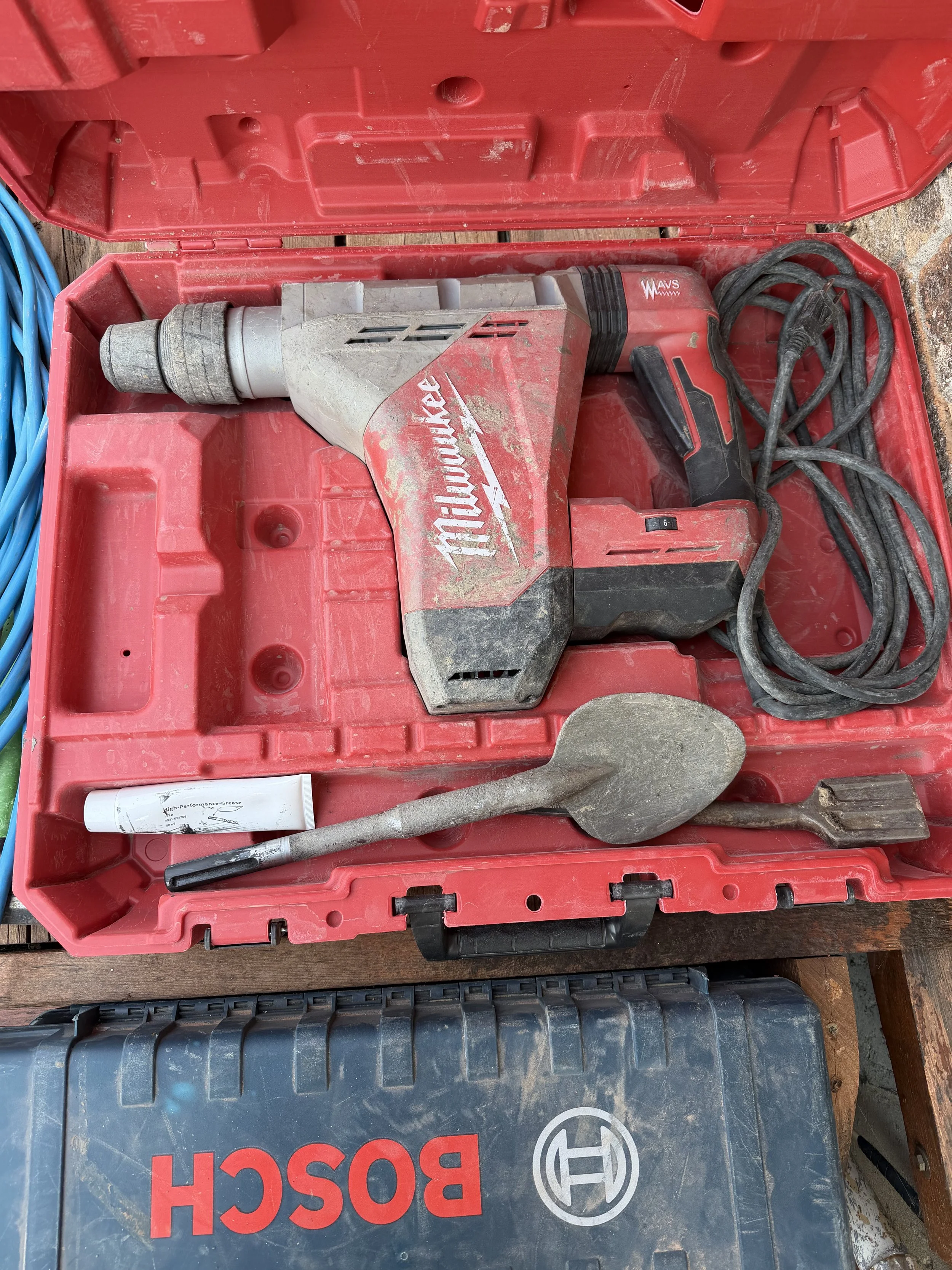 A red plastic toolbox containing a red Milwaukee cordless jack hammer, a chisel, and a tube of high-performance grease. The toolbox is open on a wooden surface with some tools nearby.