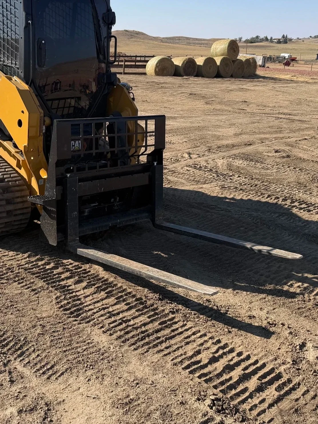 A close-up view of a forklift on a dirt surface, showing the forks extended forward and part of the forklift's front structure and attachments.