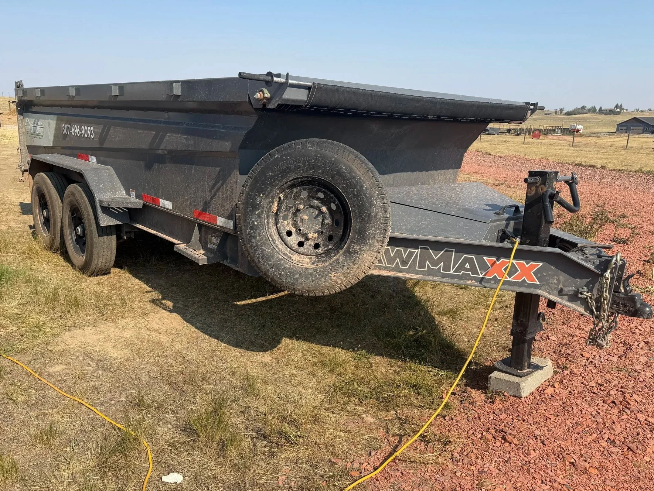 A black utility trailer with a tire mounted on its side, parked on grass and dirt in an open rural area with a few small buildings and fields in the background.
