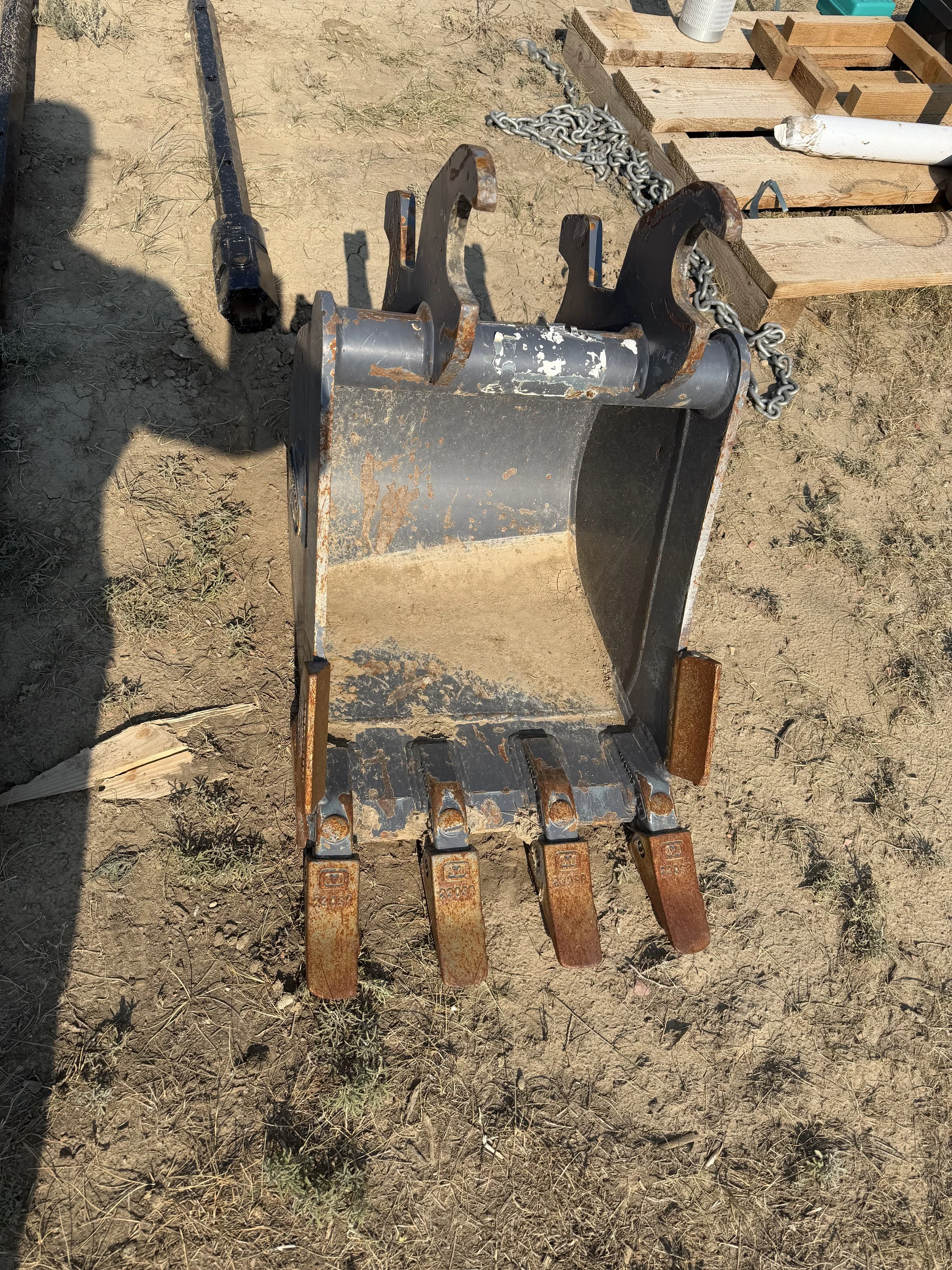 A close-up of a mechanical excavator bucket attachment with rusted teeth resting on dry dirt ground, with some wooden pallets, chains, and tools behind it.