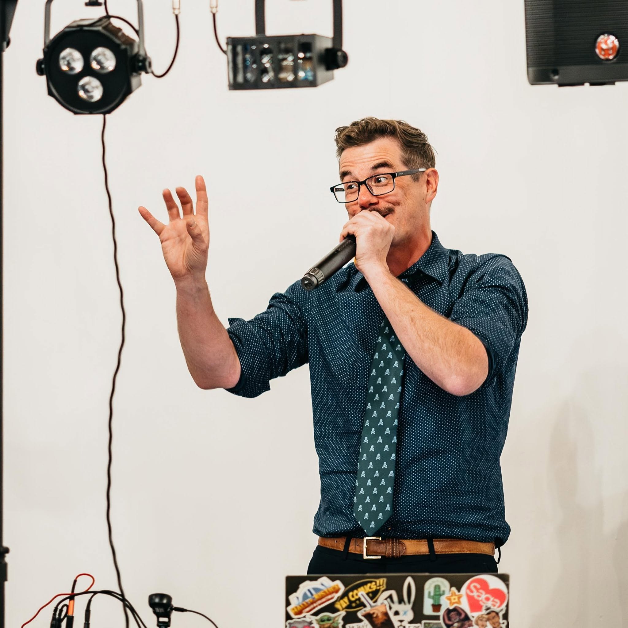 A man wearing glasses and a dark blue shirt with a pattern is speaking into a microphone, gesturing with his right hand, with a plain white wall in the background. There are some equipment and stickers partially visible in the foreground.