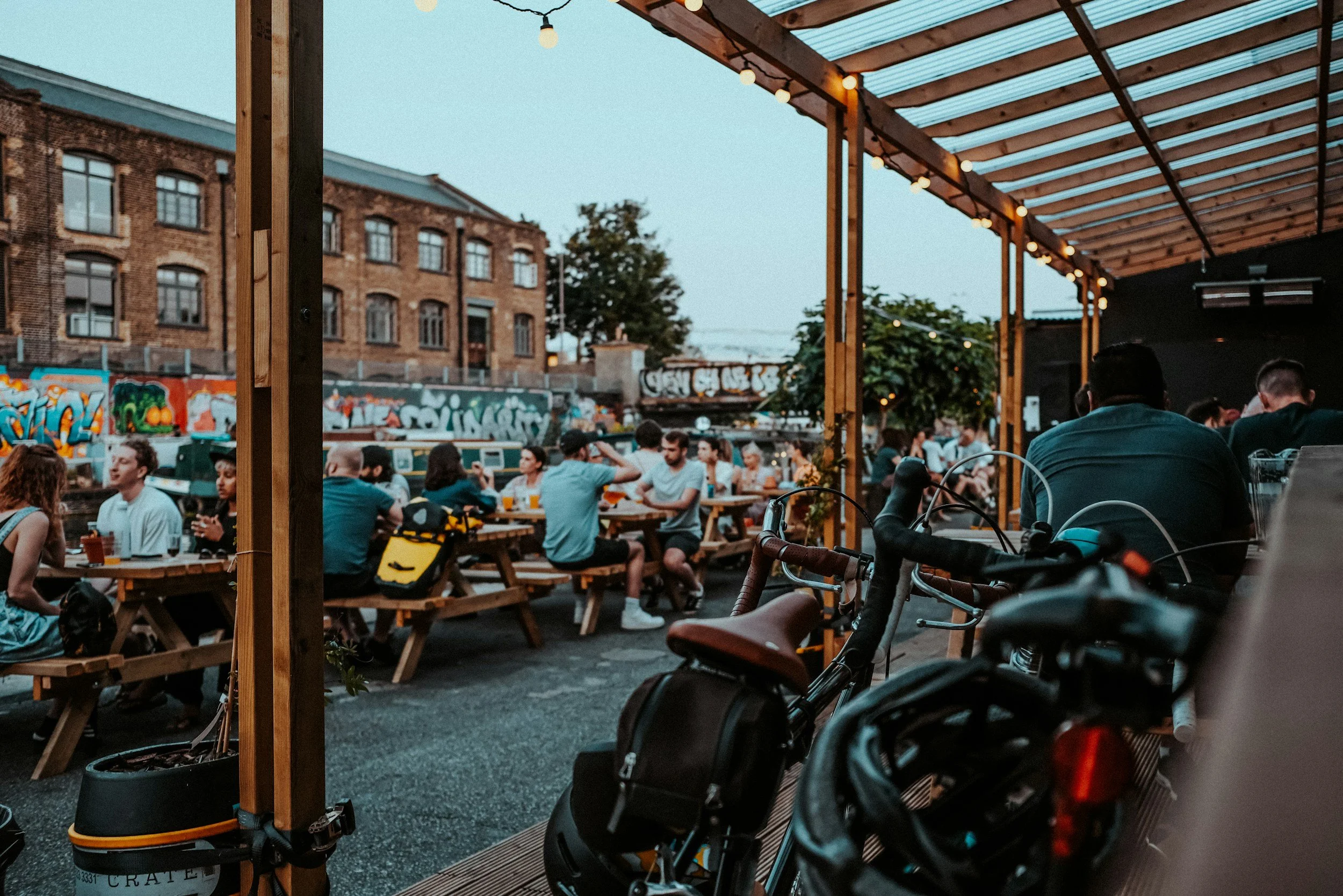 Outdoor patio with wooden tables and benches filled with people dining, string lights hanging overhead, graffiti on the brick wall in the background, and bicycles parked in the foreground during dusk.