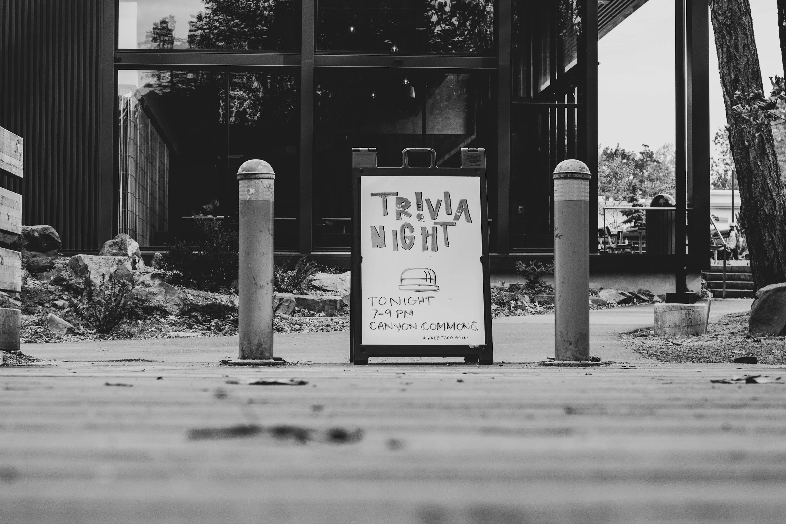 A black and white photo of a sidewalk with a signboard advertising a trivia night event at Canyon Commons from 7 to 9 PM, featuring a free taco bell. The background shows a building with large glass windows and some trees.