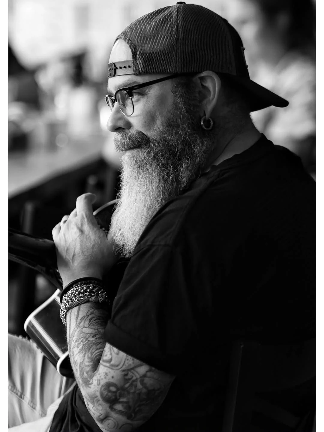 A profile view of a man with a long beard, wearing glasses, a backward baseball cap, and tattoos on his arm. He is seated indoors, holding a tablet, with a focused expression.