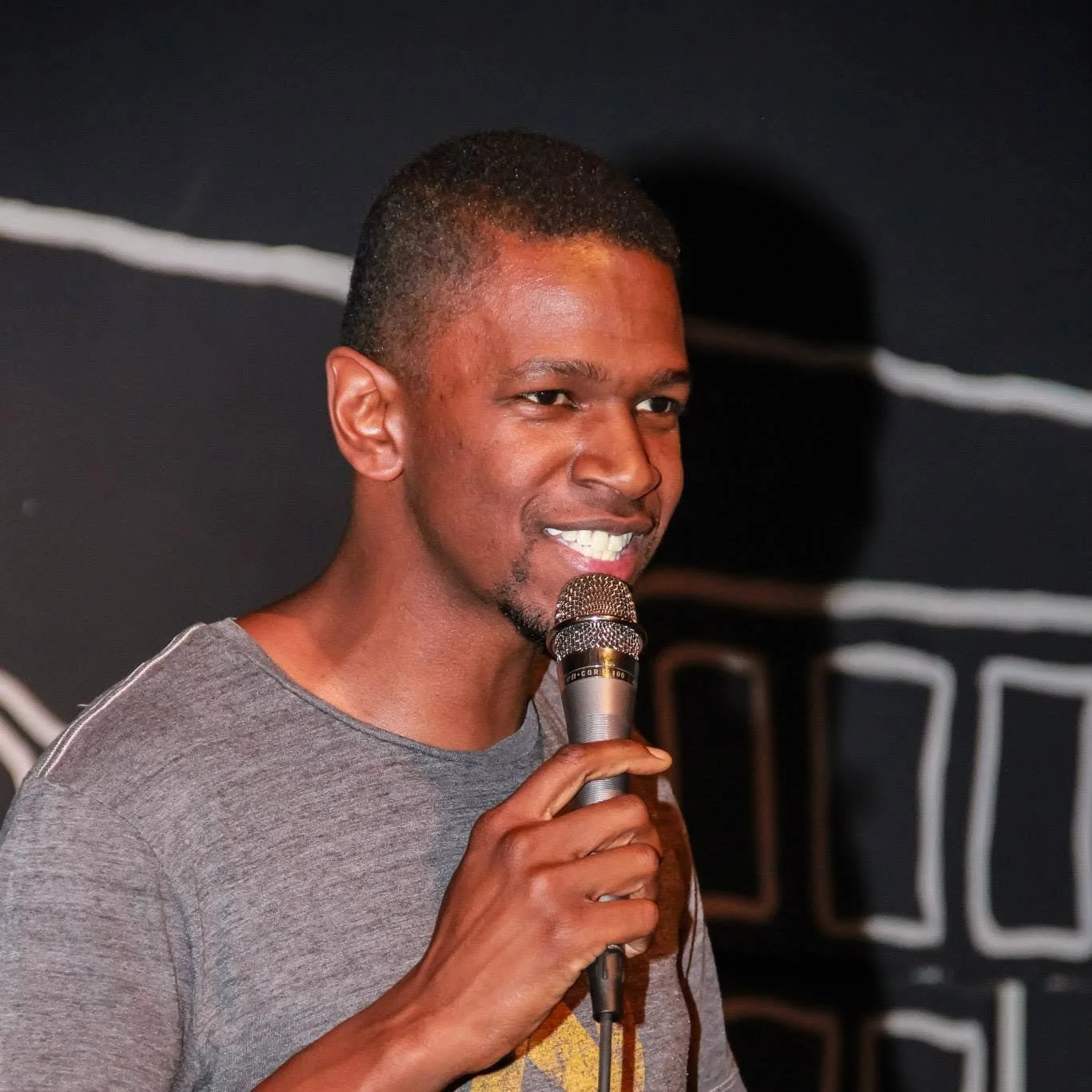 A young man speaking into a microphone on stage, wearing a gray t-shirt, with a black background behind him.