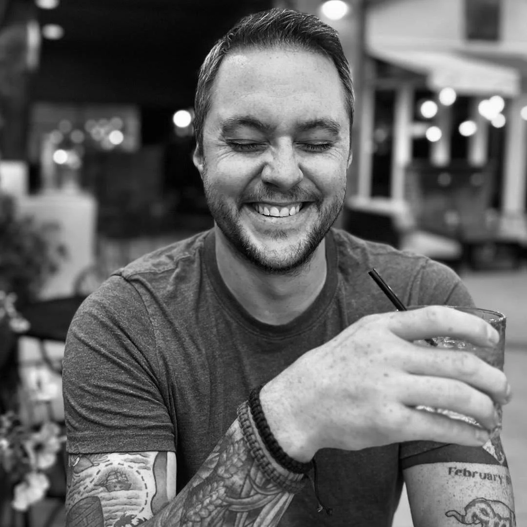 A smiling man with tattoos on his left arm holding a drink with a straw in a casual restaurant or cafe setting, in black and white.
