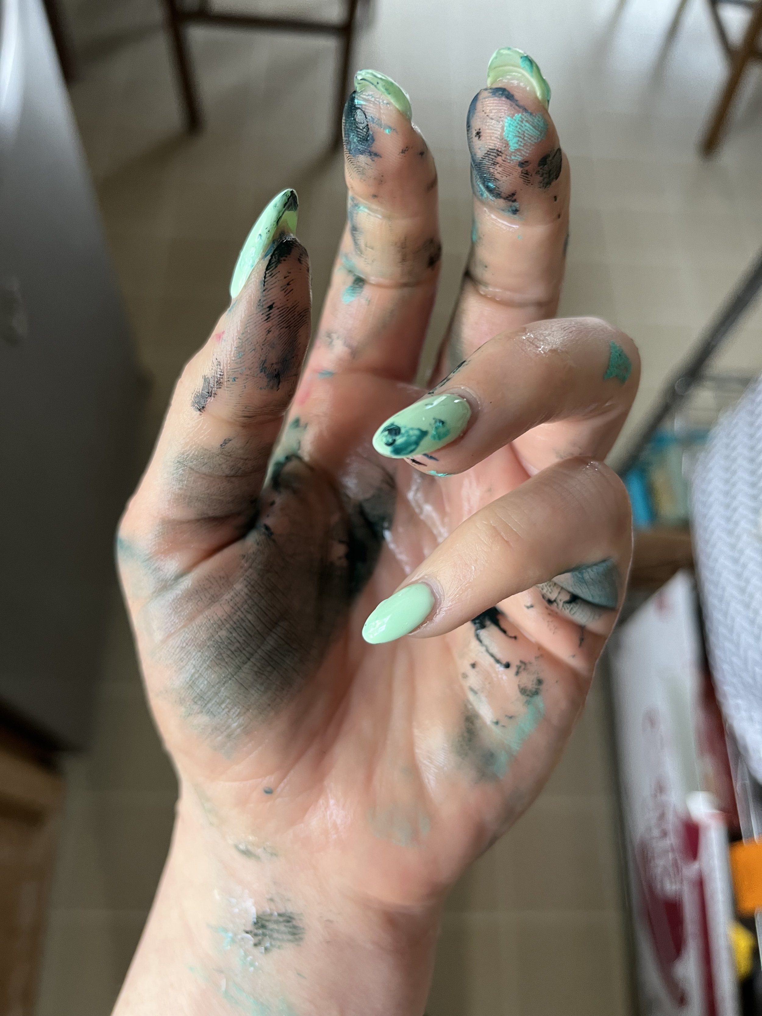 A person's painted fingers with mint green nail polish and black, blue, and pink paint smudges, suggesting finger painting or artistic activity.