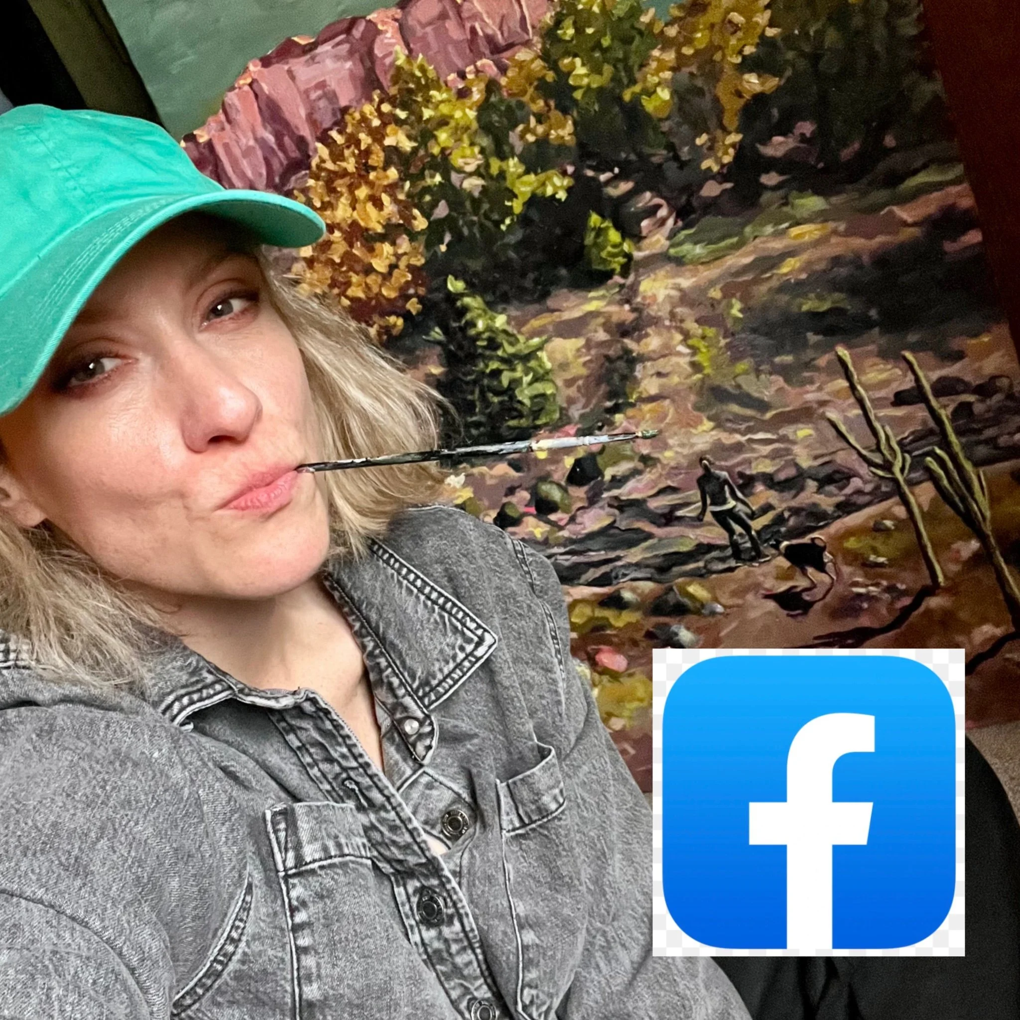 A woman with blonde hair wearing a teal baseball cap and a gray denim jacket, holding a paintbrush in her mouth, sitting in front of a colorful mountain landscape painting.