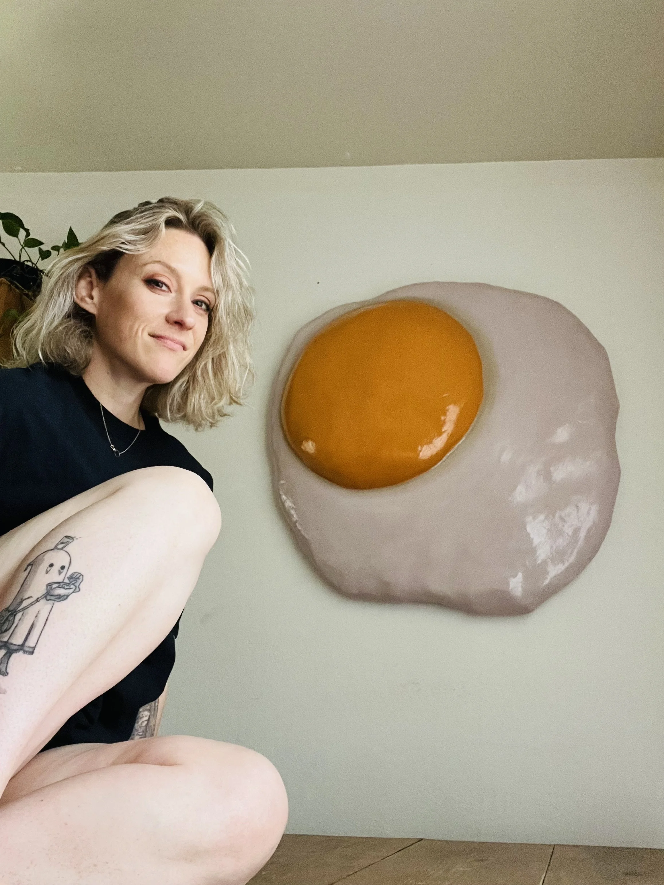 A woman with blonde hair and tattoos on her leg poses next to a wall-mounted art piece resembling a fried egg.