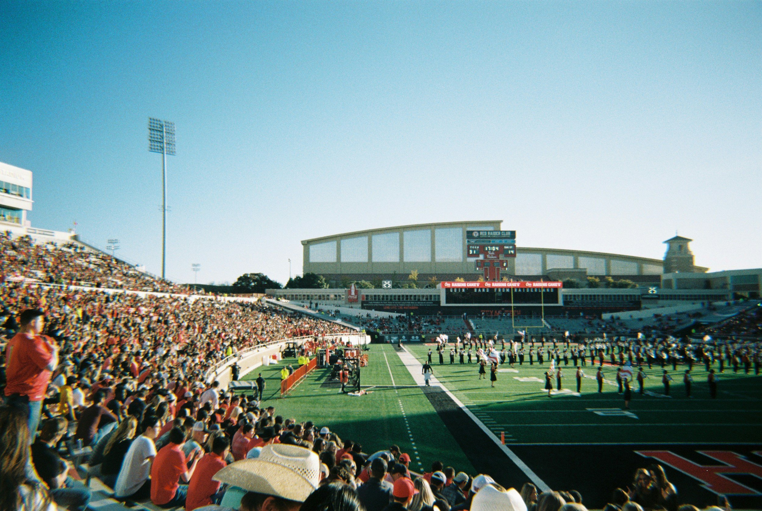 A large busy football stadium filled with spectators, players on the field, and a scoreboard displaying game information, under a clear blue sky.