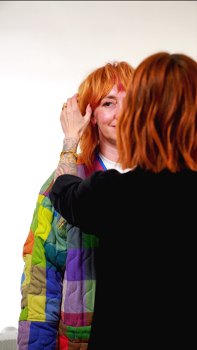 A woman with red hair smiling as another woman touches her face in an art gallery or studio setting.