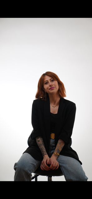 A woman with shoulder-length red hair sitting on a stool against a plain white background, wearing a black jacket and jeans.