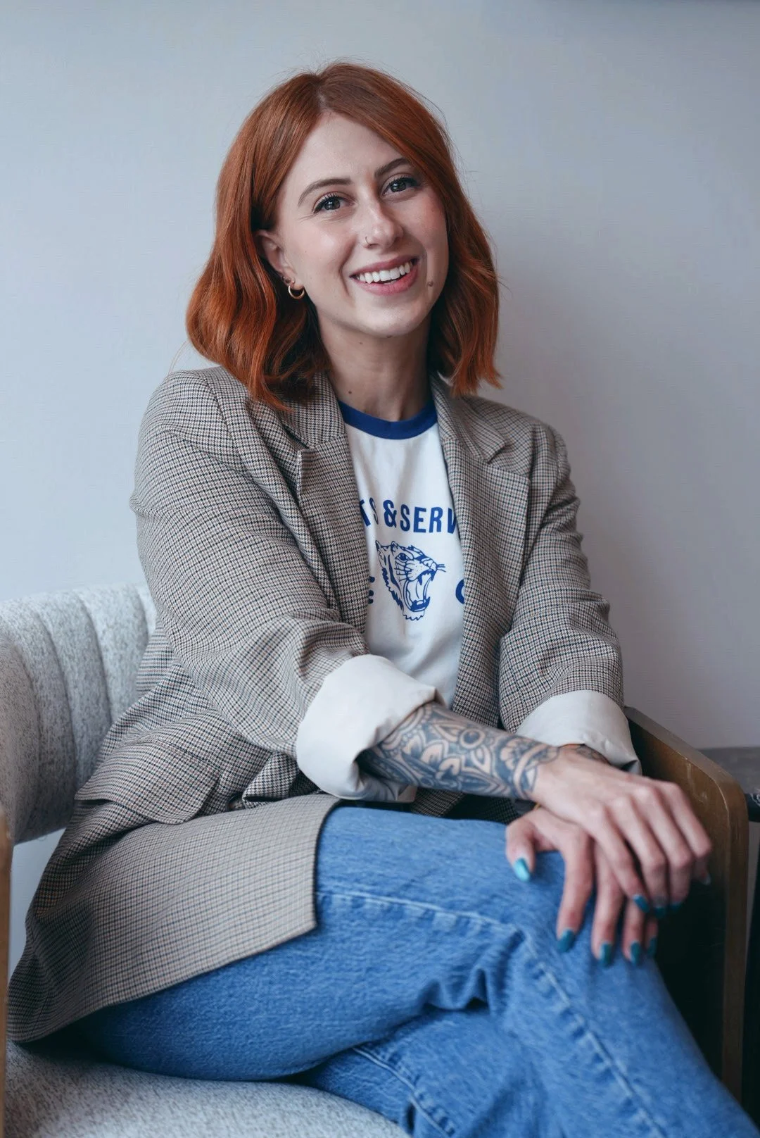 A woman with red hair and tattoos on her right arm, wearing a checked blazer and jeans, sitting on a beige upholstered chair, smiling at the camera.