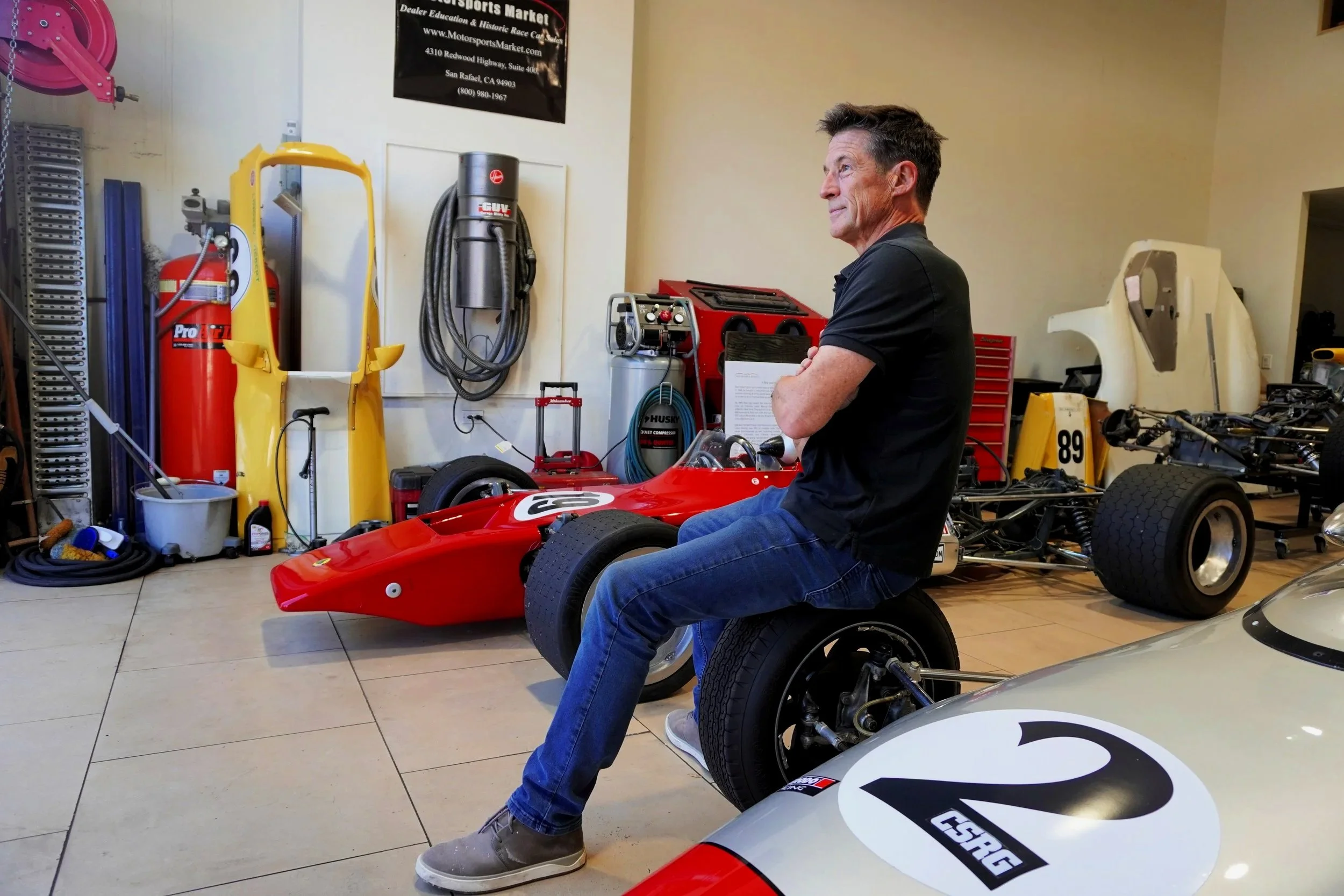A man with short hair and a black shirt sitting in a race car in a garage or workshop. The garage has various tools and equipment, including a fire extinguisher, hoses, a toolbox, and a partially disassembled race car frame. There is a black banner on the wall with white and red text and a website URL.