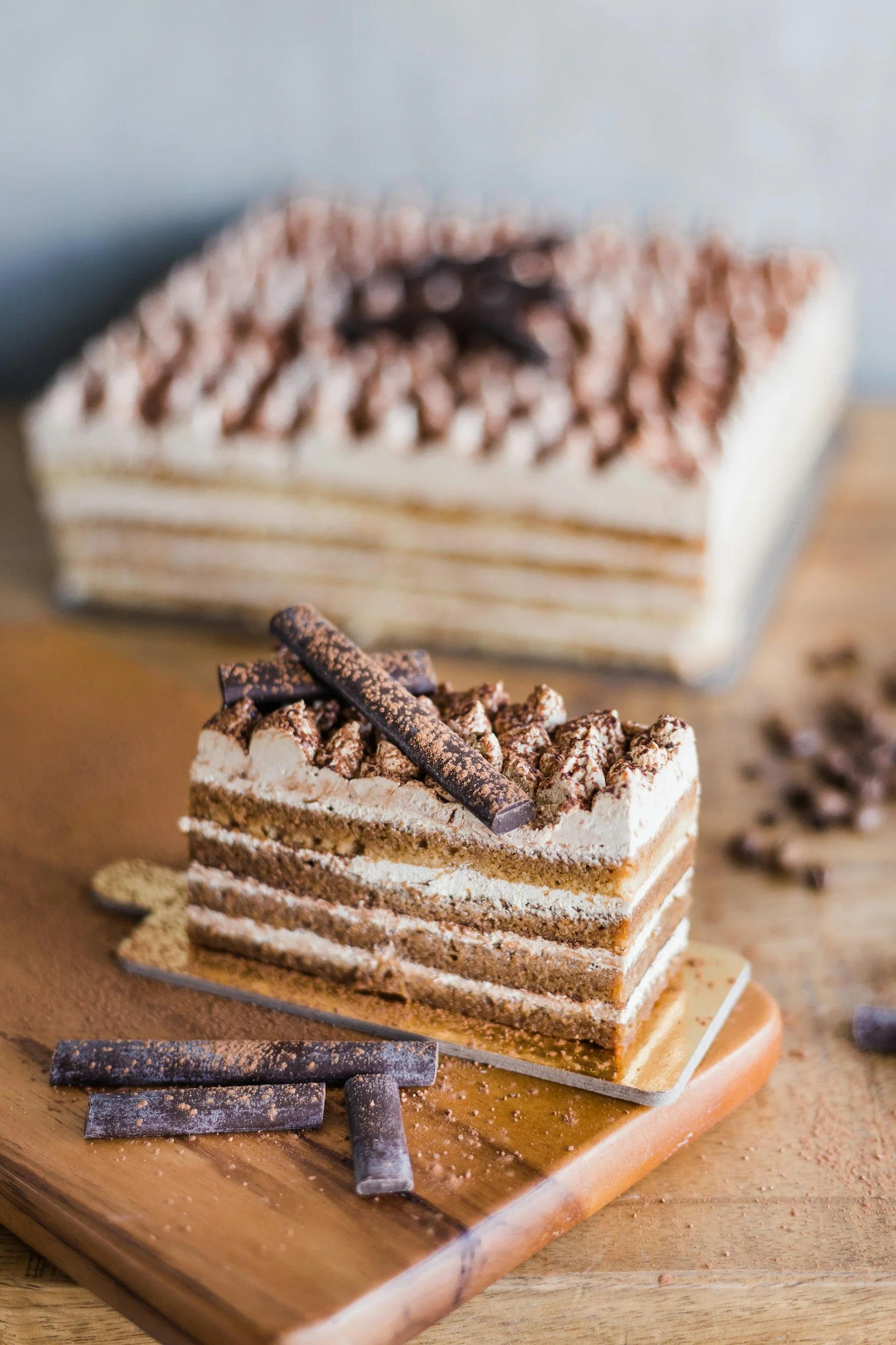 A slice of layered chocolate and vanilla cake topped with chocolate shavings and powdered cocoa on a wooden serving board, with additional chocolate pieces in the background.