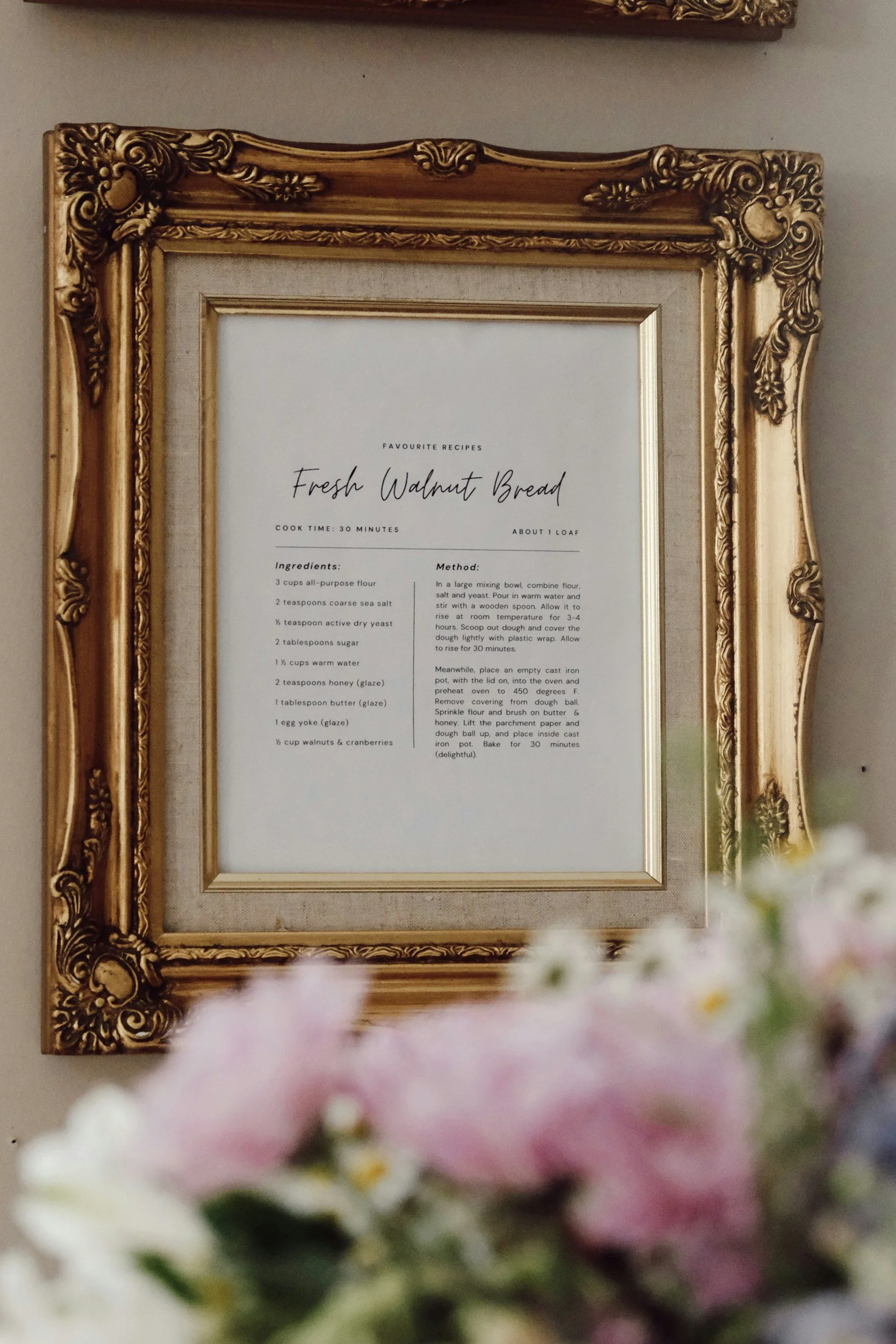 Framed recipe for Fresh Walnut Bread on a beige wall, surrounded by pink and white flowers in the foreground.