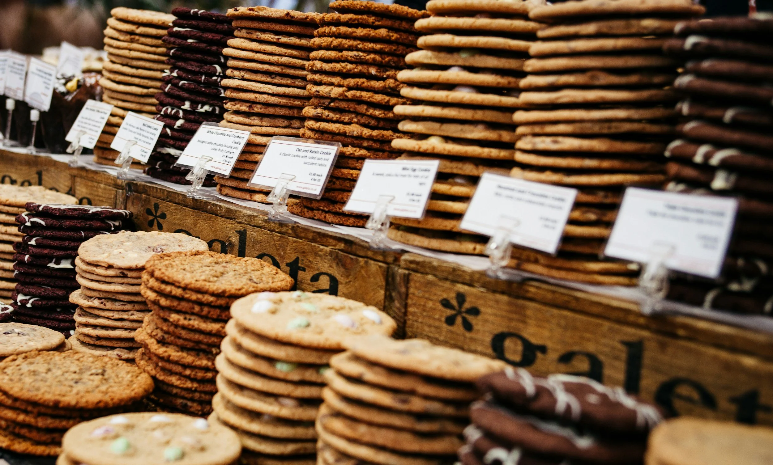 Stacks of assorted cookies, including chocolate and oatmeal cookies, displayed on a wooden stand at a market or bakery.