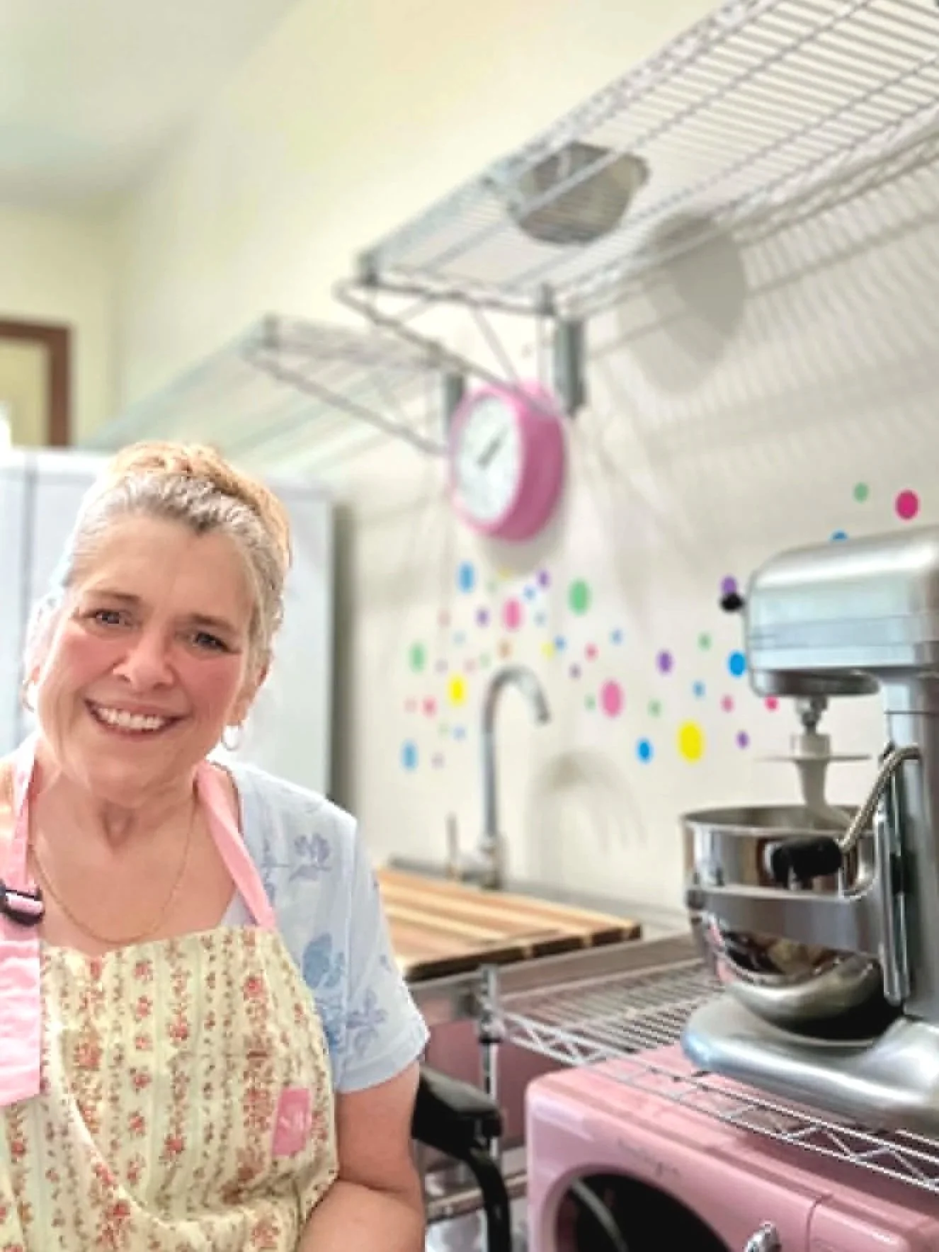 Smiling woman in apron in a colorful kitchen with a stand mixer, pink clock, and decorative wall dots.