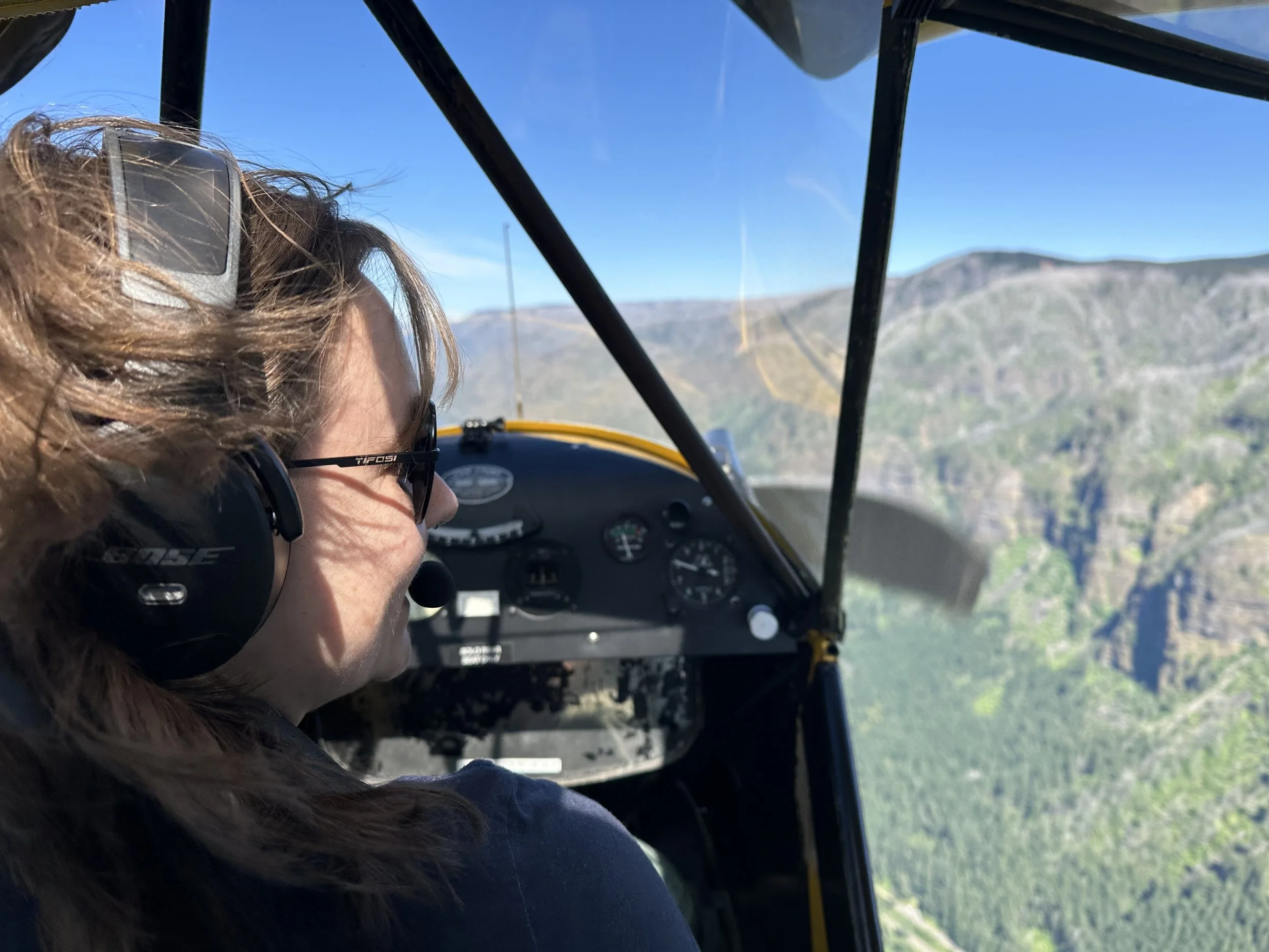 Person with curly brown hair wearing sunglasses and a headset flying in an airplane cockpit with mountainous landscape visible outside the window.