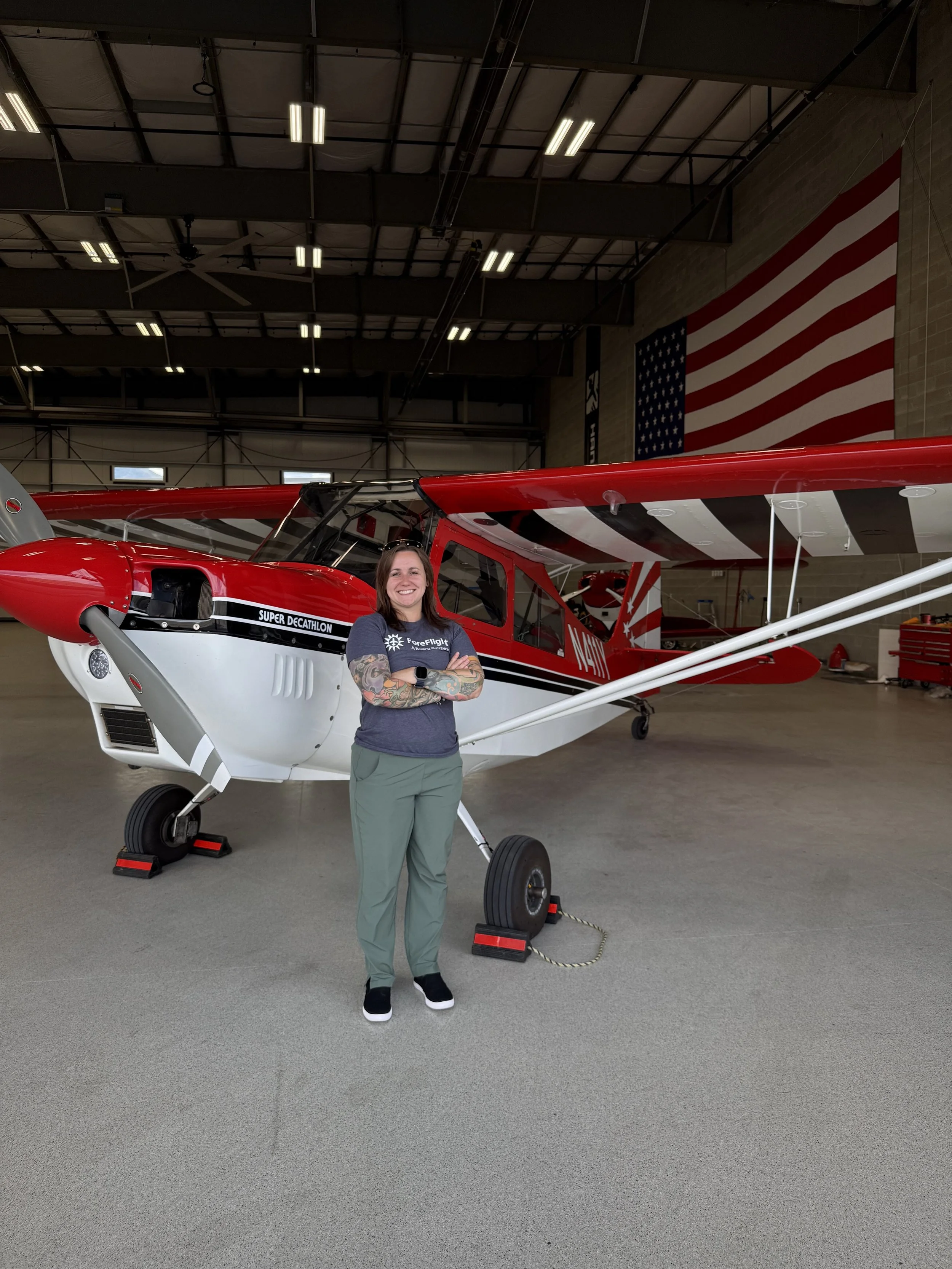 A woman smiling in front of a red and white helicopter inside a hangar, with American flag on the wall in the background.