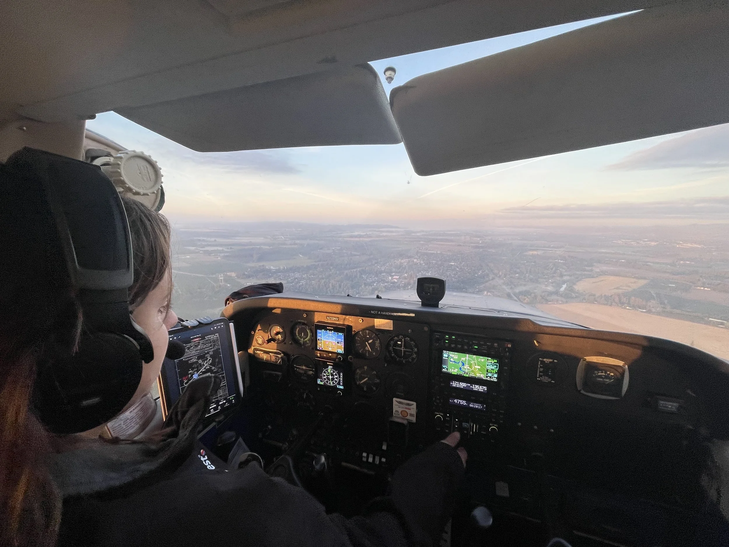 Inside the cockpit of an airplane flying above the landscape at sunset, with pilots and digital displays visible.