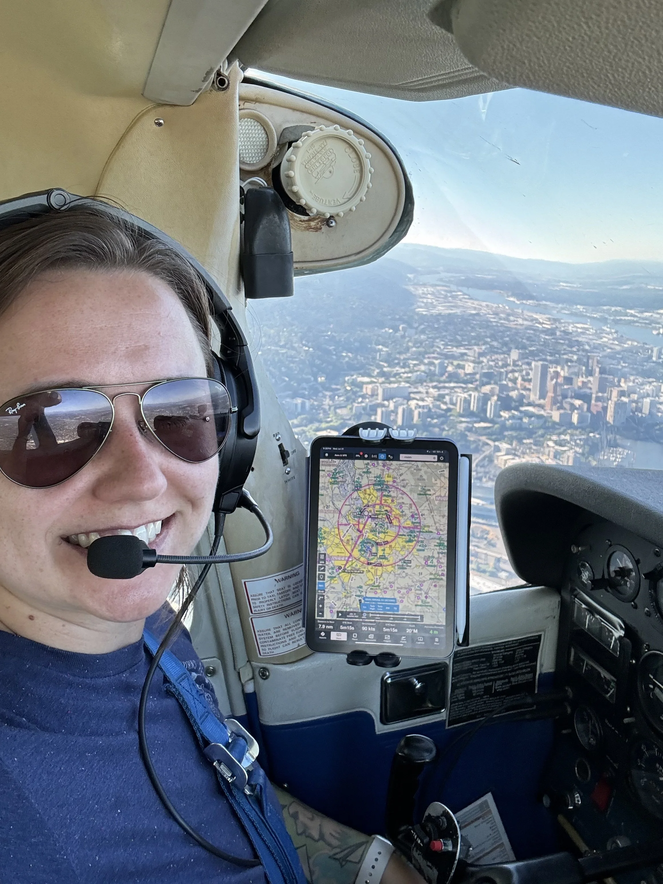 Person wearing sunglasses and a headset, sitting in the cockpit of an airplane, with a large view of a cityscape and countryside through the window; airplane instruments and a tablet with navigation data are visible.