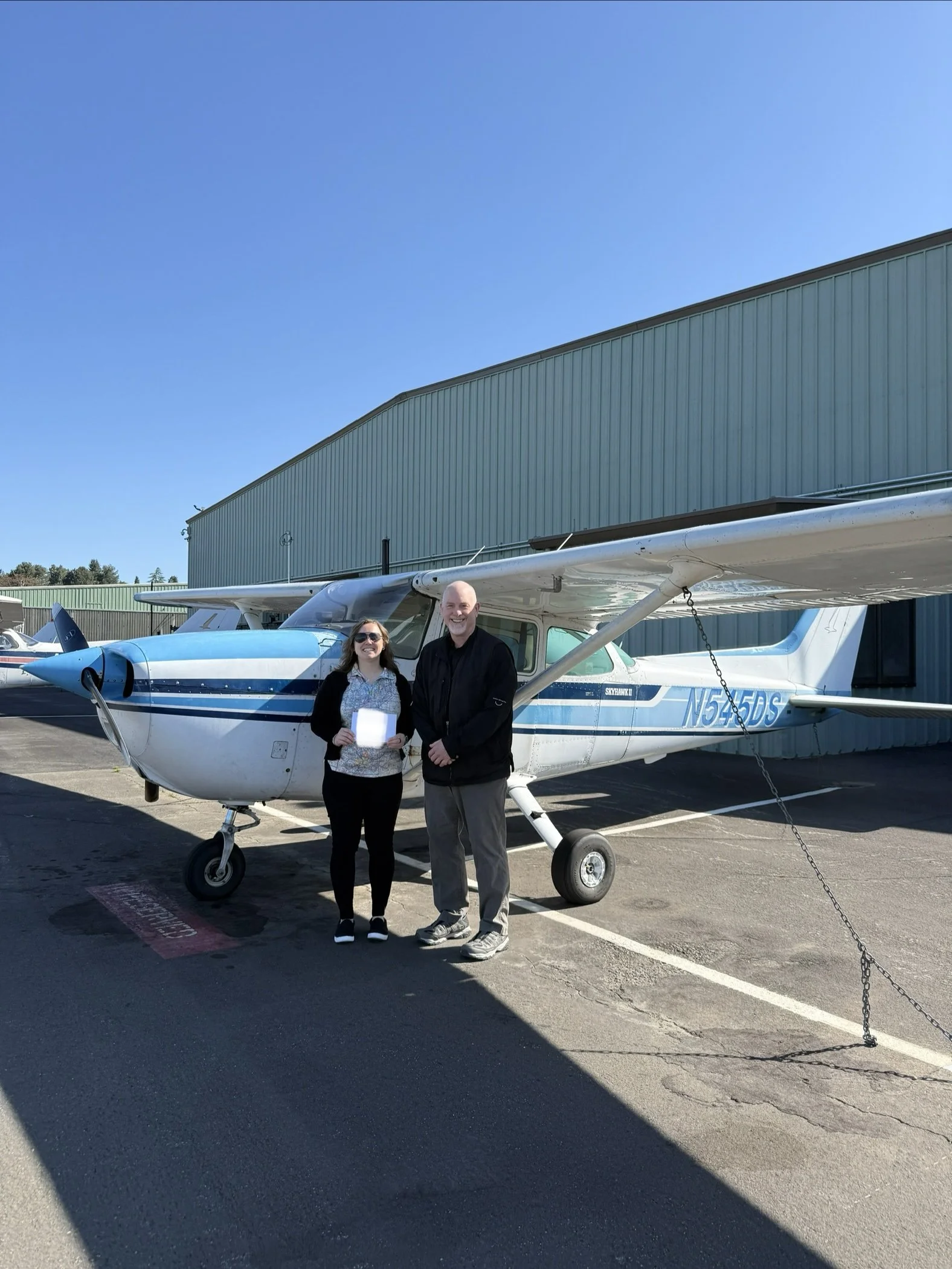 Two people standing in front of a small airplane at an airport, with a hangar in the background and clear blue sky overhead.