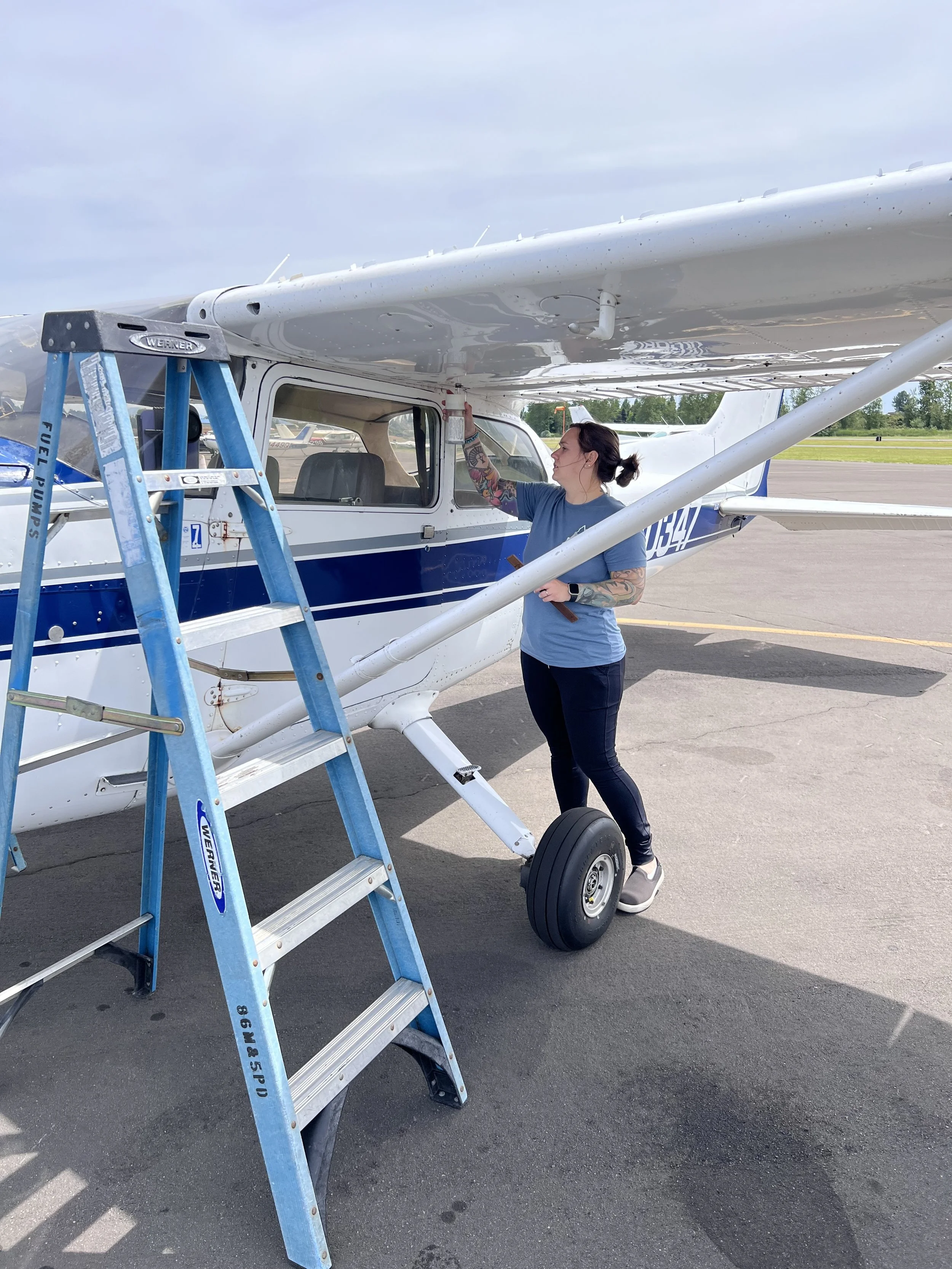 A woman with tattoos on her arm cleaning the underside of a small white airplane on an airport tarmac with a step ladder nearby.