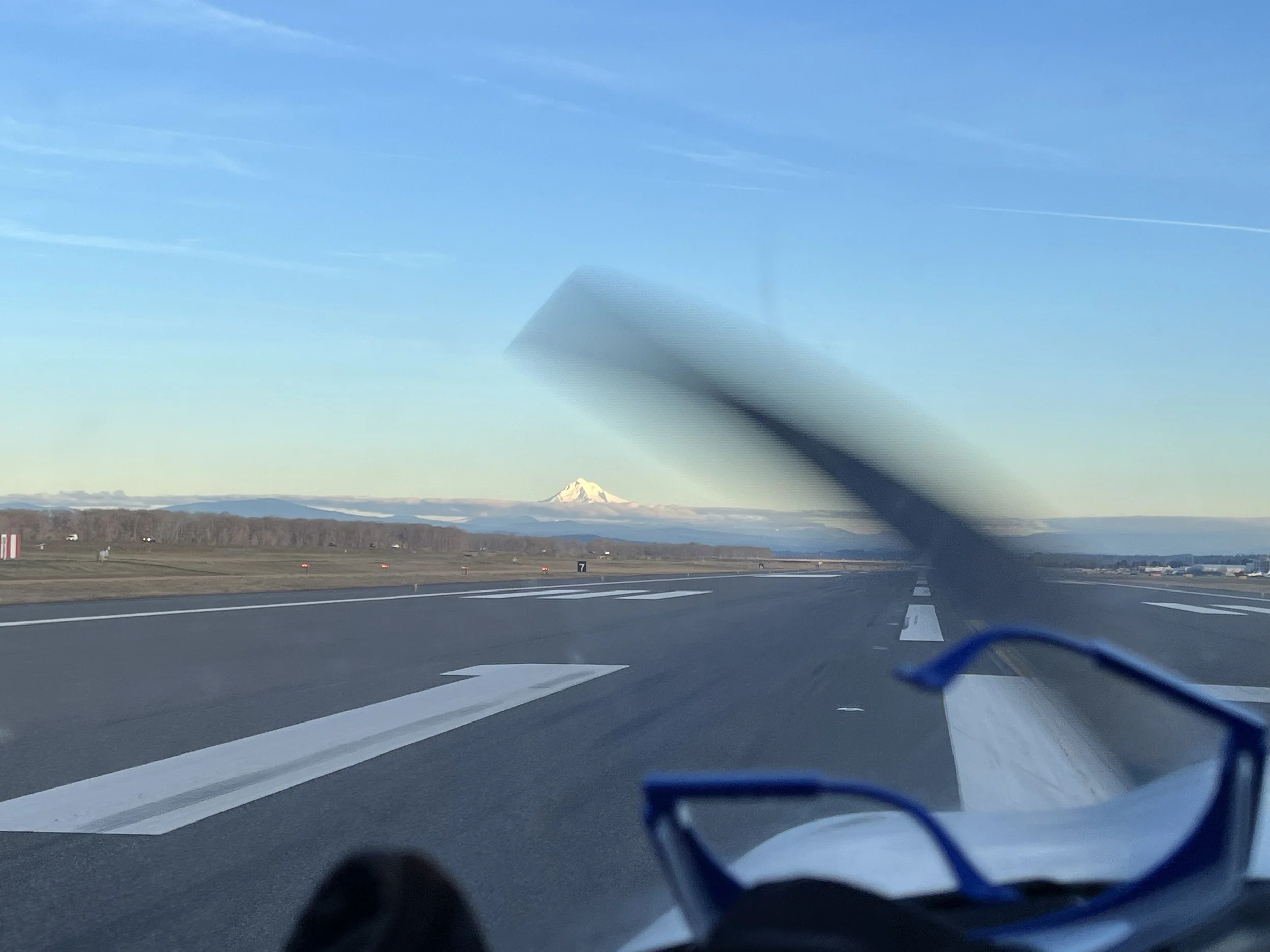 View of a highway with mountains in the distance and a clear blue sky, taken from inside a vehicle.