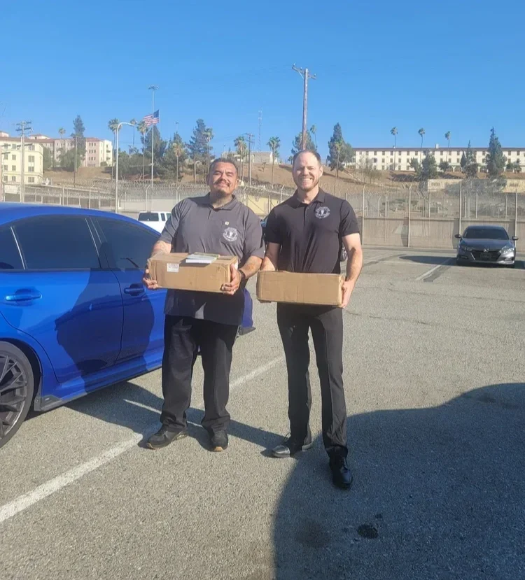 Two men in dark shirts standing in a parking lot holding cardboard boxes, with a blue car on the left and a few other cars in the background, under a clear blue sky.