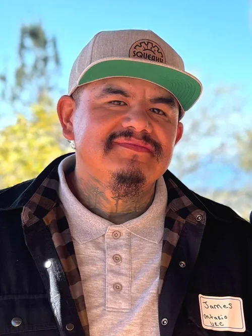 A man with a mustache and goatee wearing a gray baseball cap with a logo, a gray polo shirt, and a checkered shirt underneath. He has a name tag saying "James Ontario UBC" and is outdoors with a clear blue sky and trees in the background.