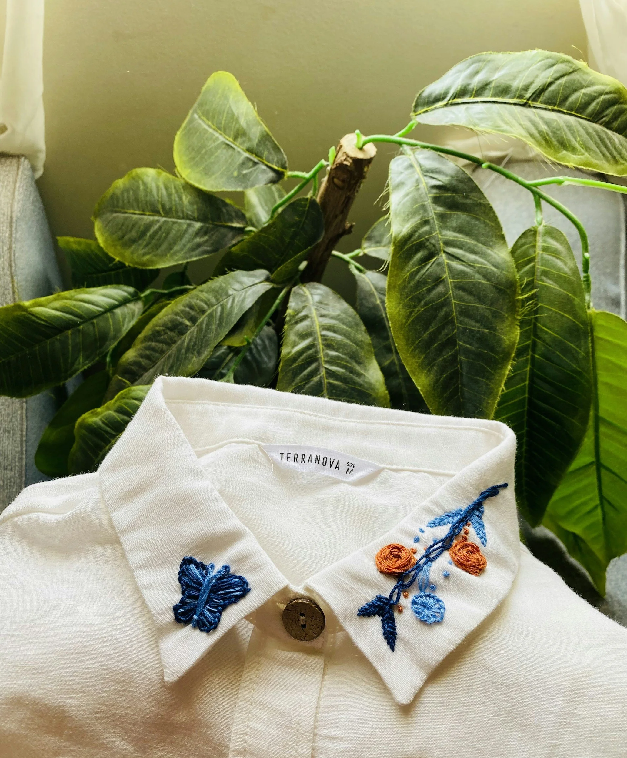 Close-up of a white shirt with embroidered butterflies and flowers, displayed in front of green houseplant leaves.