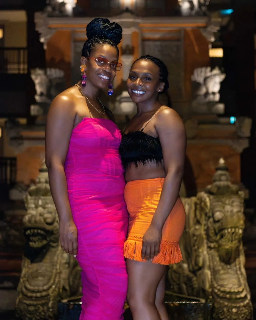 Two women smiling and embracing in front of a traditional ornate background, dressed in colorful outfits; one in a pink strapless dress and the other in a black top with an orange skirt.