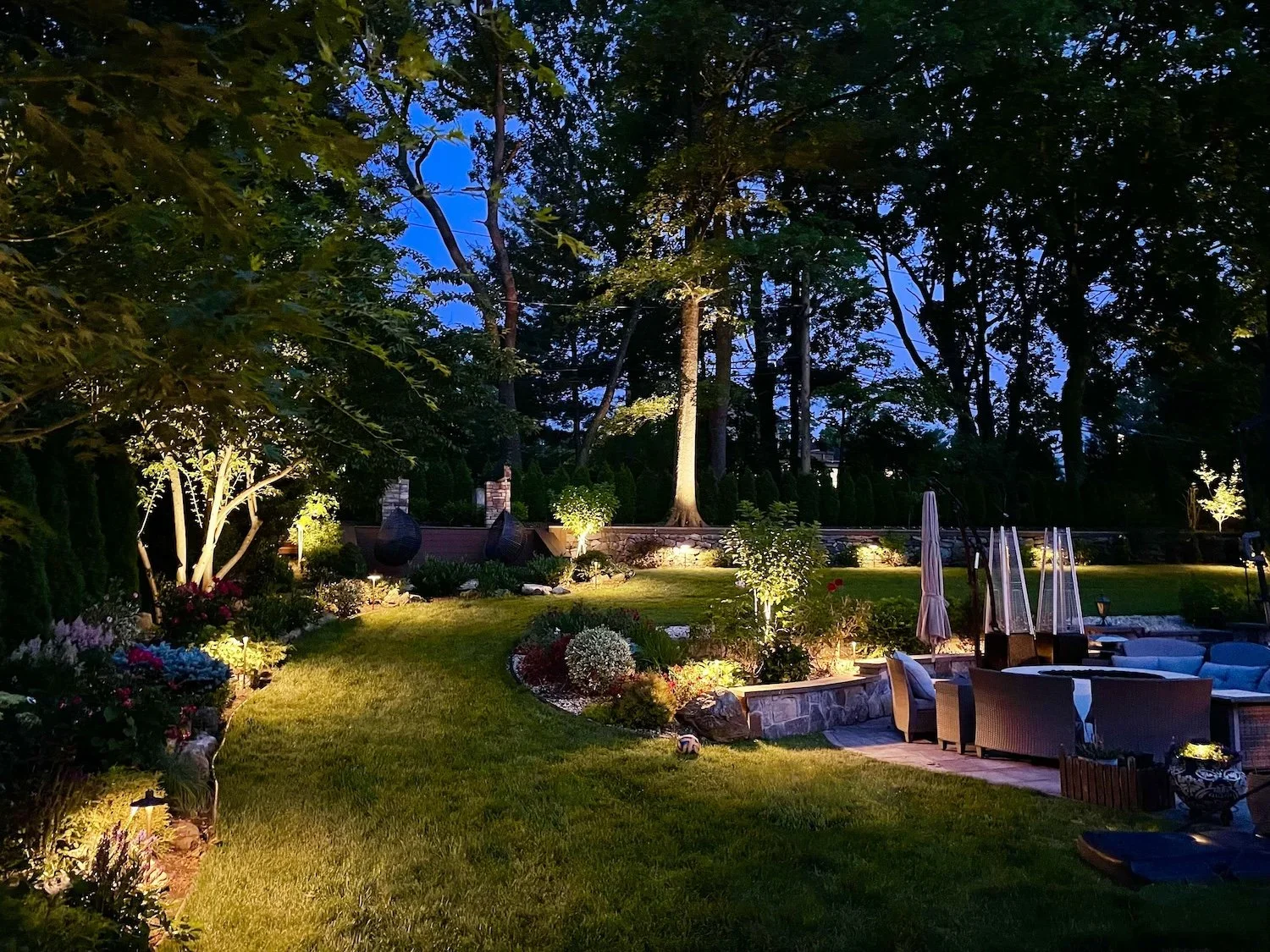 A beautifully lit backyard garden scene at dusk with trees, shrubs, flowers, outdoor furniture, and a firepit area.