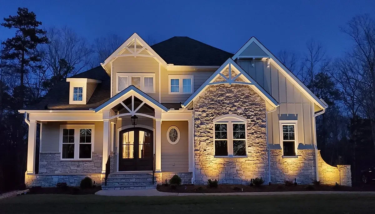 A large, modern house illuminated at dusk with exterior lighting, featuring stone and siding construction, multiple gables, and a staircase leading to a front door surrounded by a well-maintained lawn.