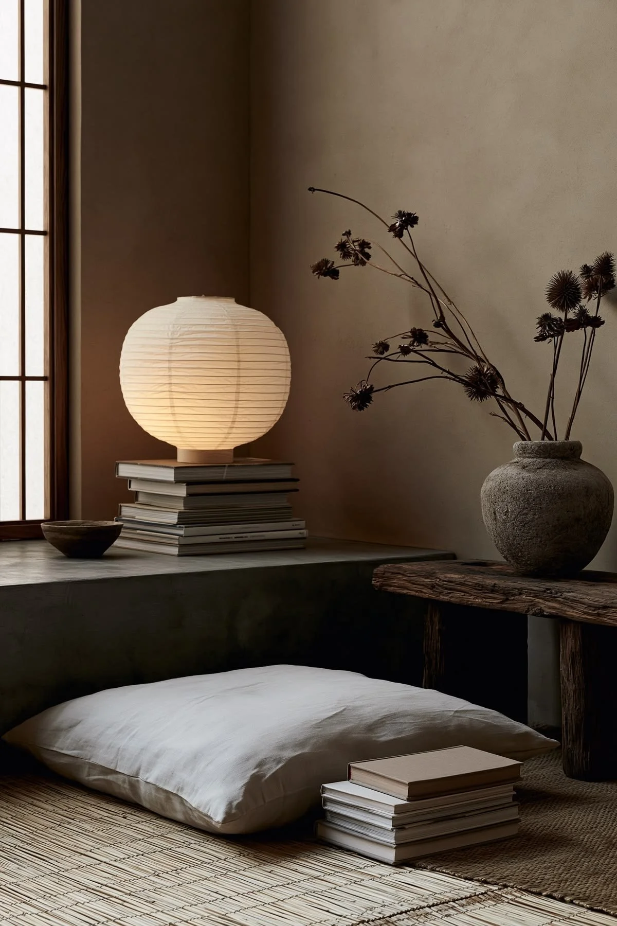 Minimalist bedroom scene with a lit paper lantern on top of stacked books next to a ceramic vase with dried flowers, a bowl, a mattress with a pillow, and a rolled-up mat, all near a shoji screen window.