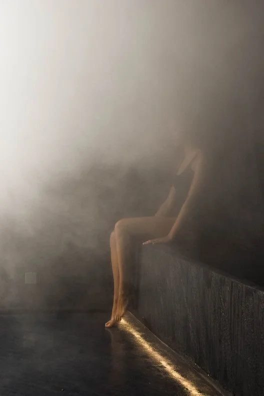 A woman in a black dress sitting on the edge of a dark platform, with fog or smoke surrounding her, in a dimly lit room.