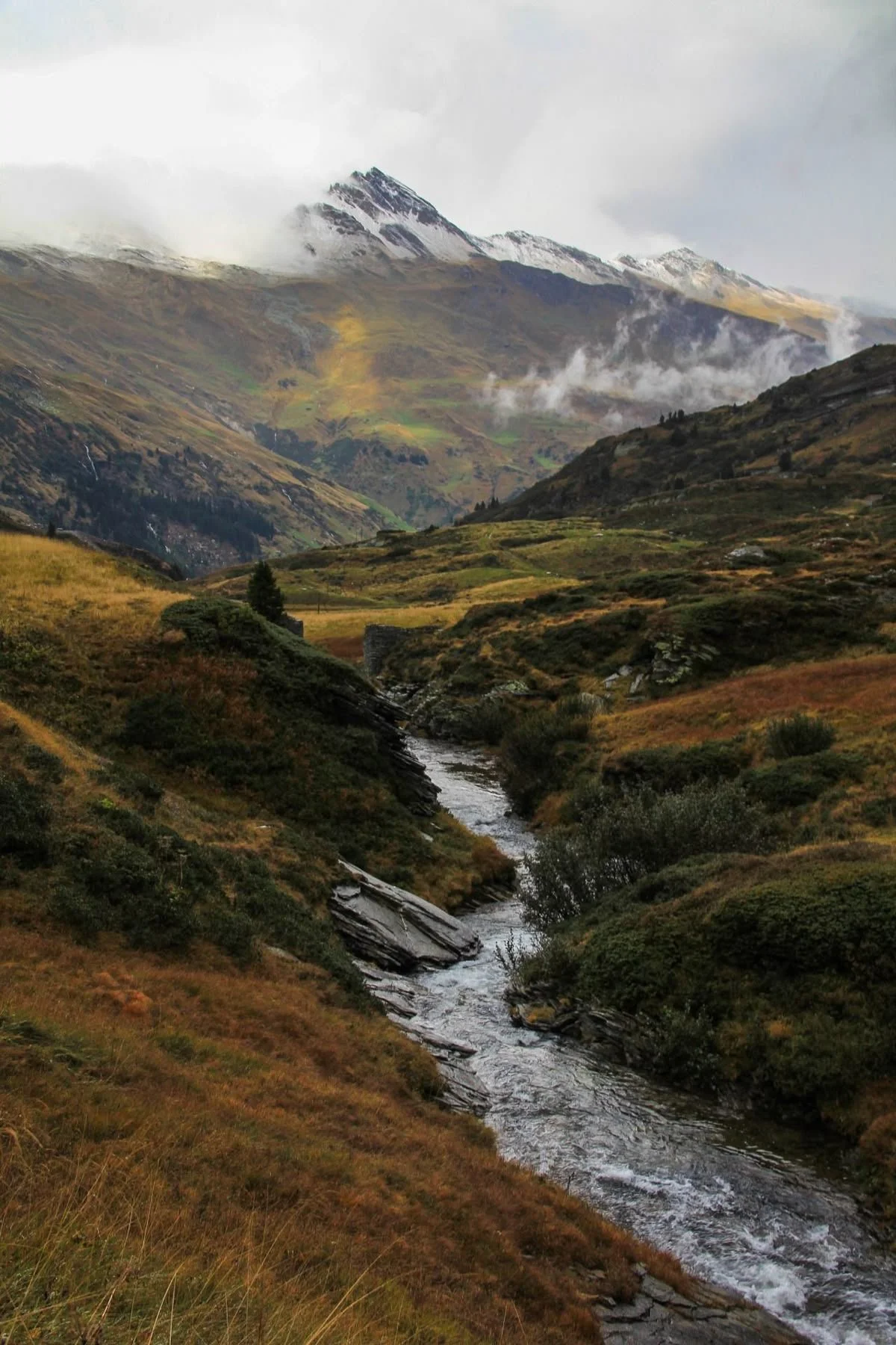 A mountain landscape with a river running through lush green hills, snow-capped peaks in the background, and mist or clouds around the mountain tops.