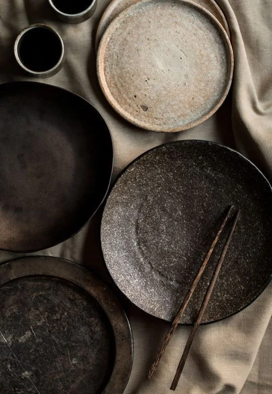 Several black and grey ceramic plates and bowls, with wooden chopsticks resting on one plate, arranged on a grey fabric surface.