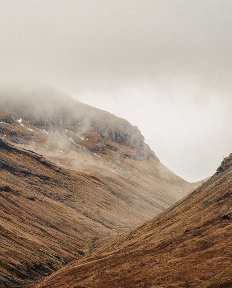 A mountain valley with steep, brown, grassy slopes and mist-covered peaks in the background.