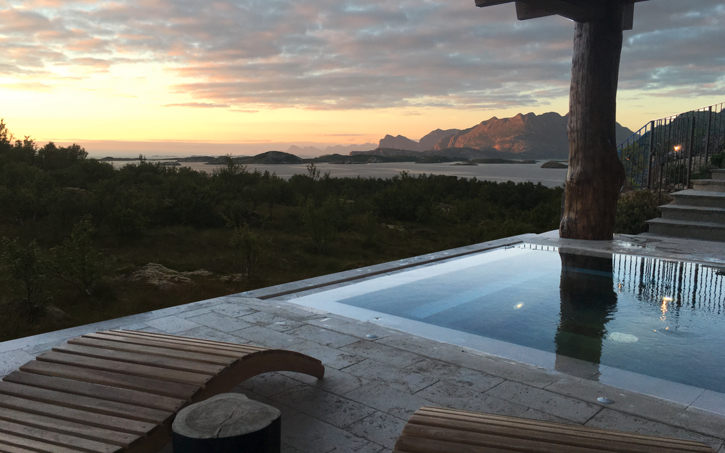 View of a sunset over a mountainous landscape with water, seen from a patio with wooden lounge chairs, a hot tub, and a tree.