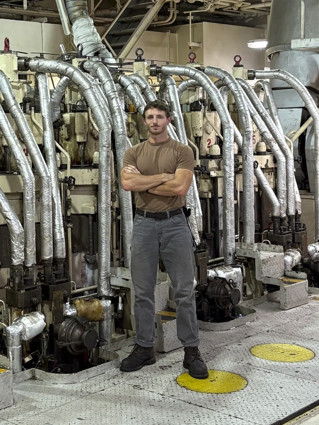 A man standing with crossed arms in front of a complex array of metallic pipes and industrial machinery in a factory or plant setting.