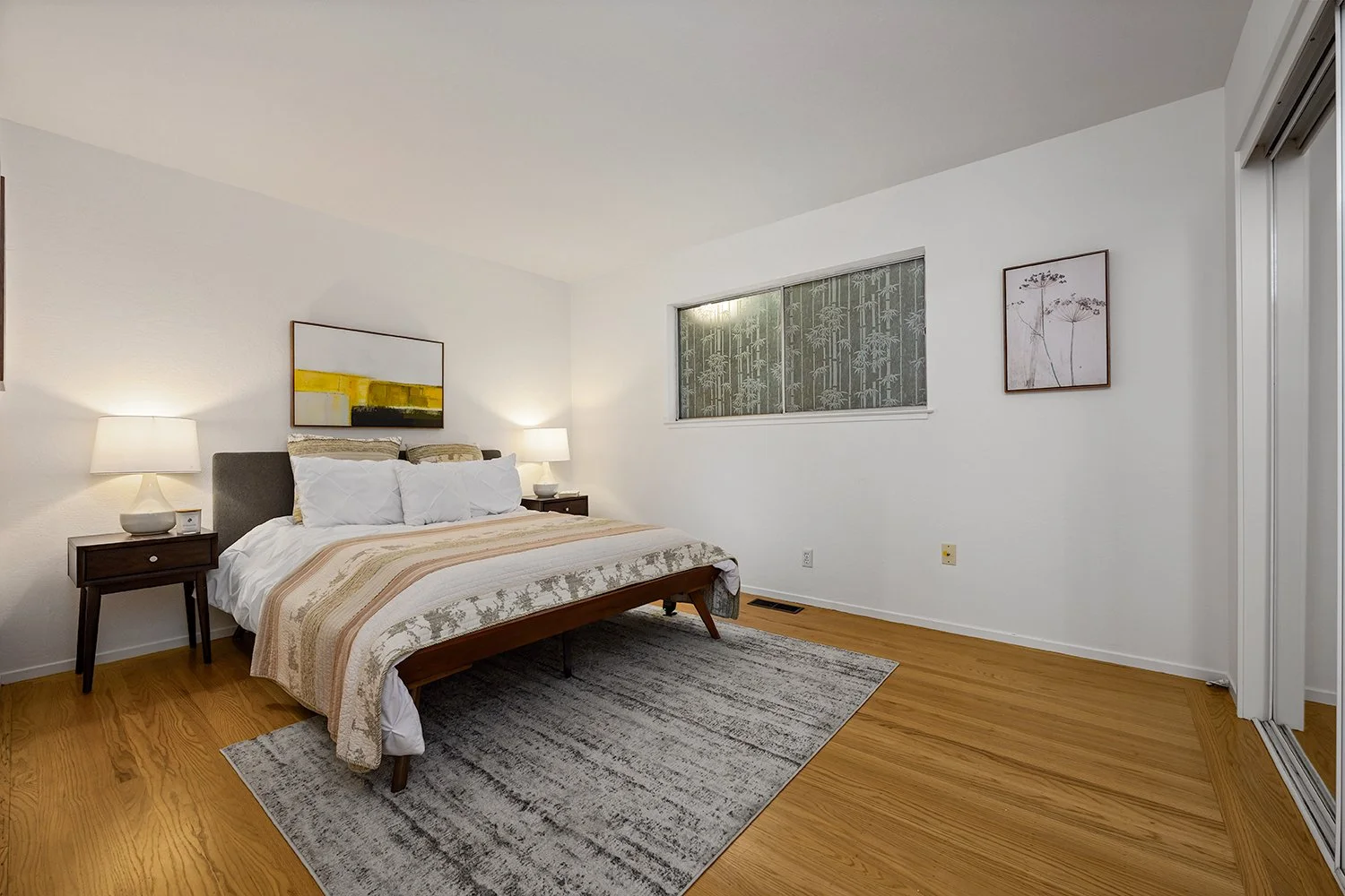 A bedroom with a bed, two table lamps, and minimal artwork on white walls. Wooden floors and a window with patterned curtains.