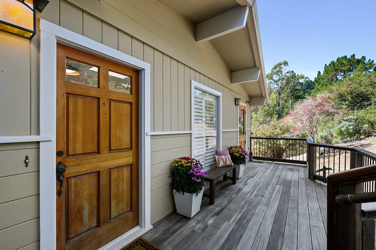 Front porch of a house with a wooden door, potted flowers, a bench with cushions, and a scenic view of trees and plants.