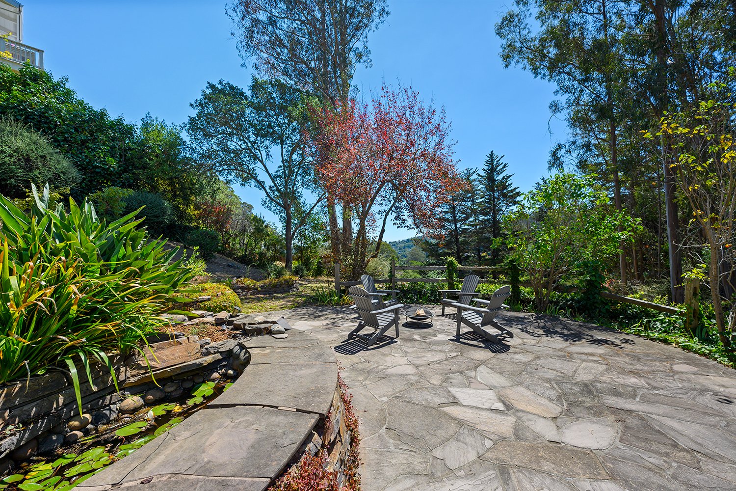 Outdoor patio with four Adirondack chairs surrounding a fire pit, surrounded by lush green trees and plants, under a bright blue sky.