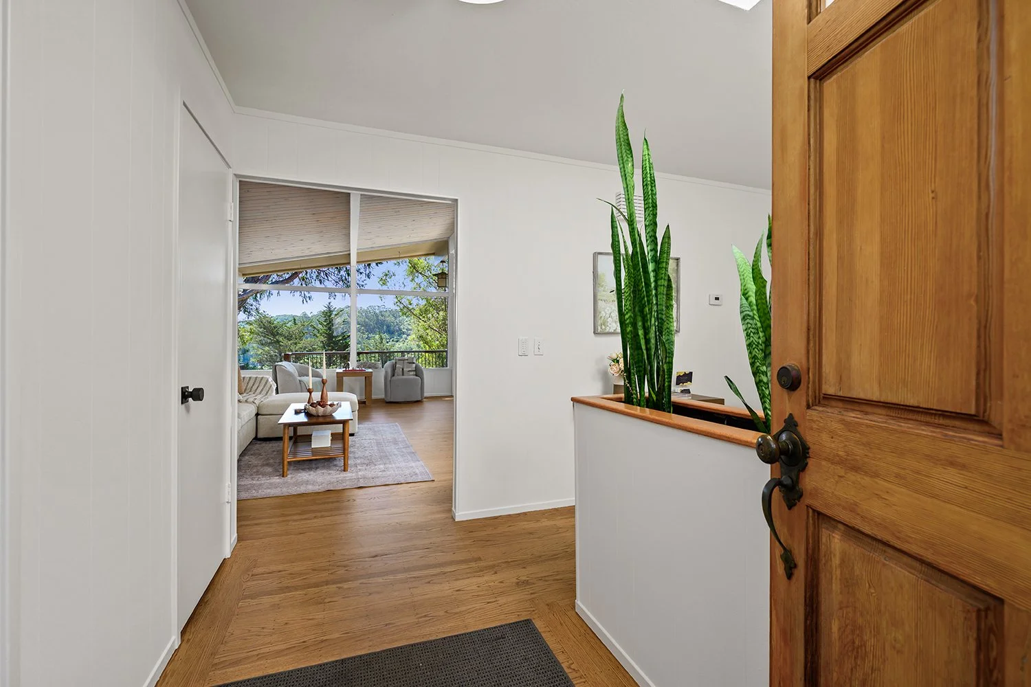 Entryway with a wooden door open to a living room with large windows, white walls, wooden floors, and modern furniture, including a white couch and a small table with decorative items.