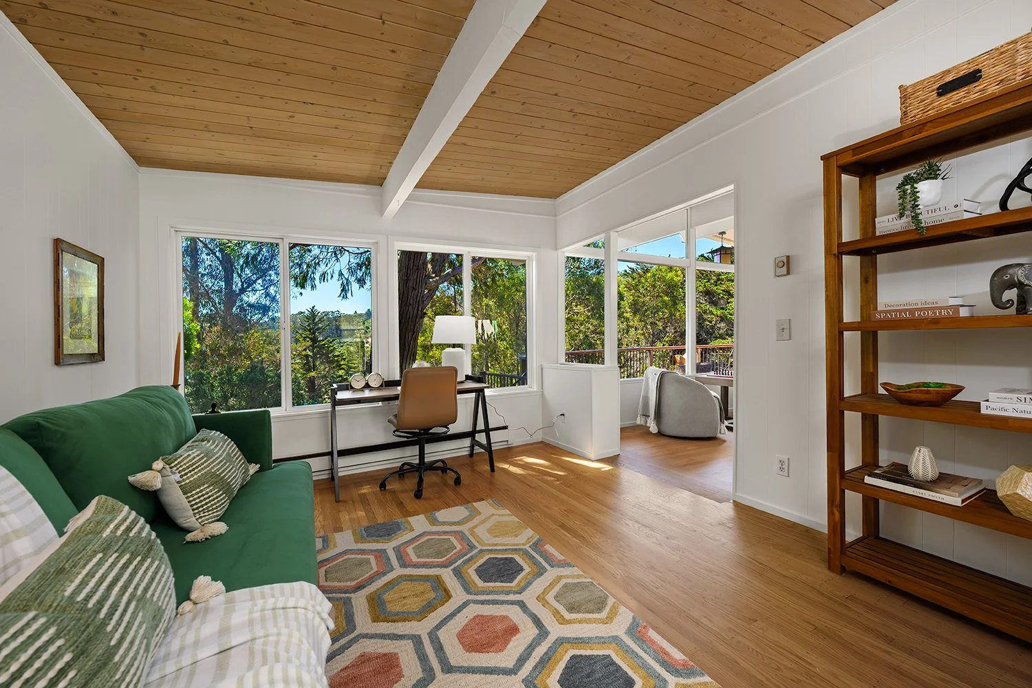 Bright living room with large windows showing outdoor trees, including a green sofa with patterned pillows, a geometric area rug, a small desk with a beige chair, and a wooden bookshelf with decorative items.