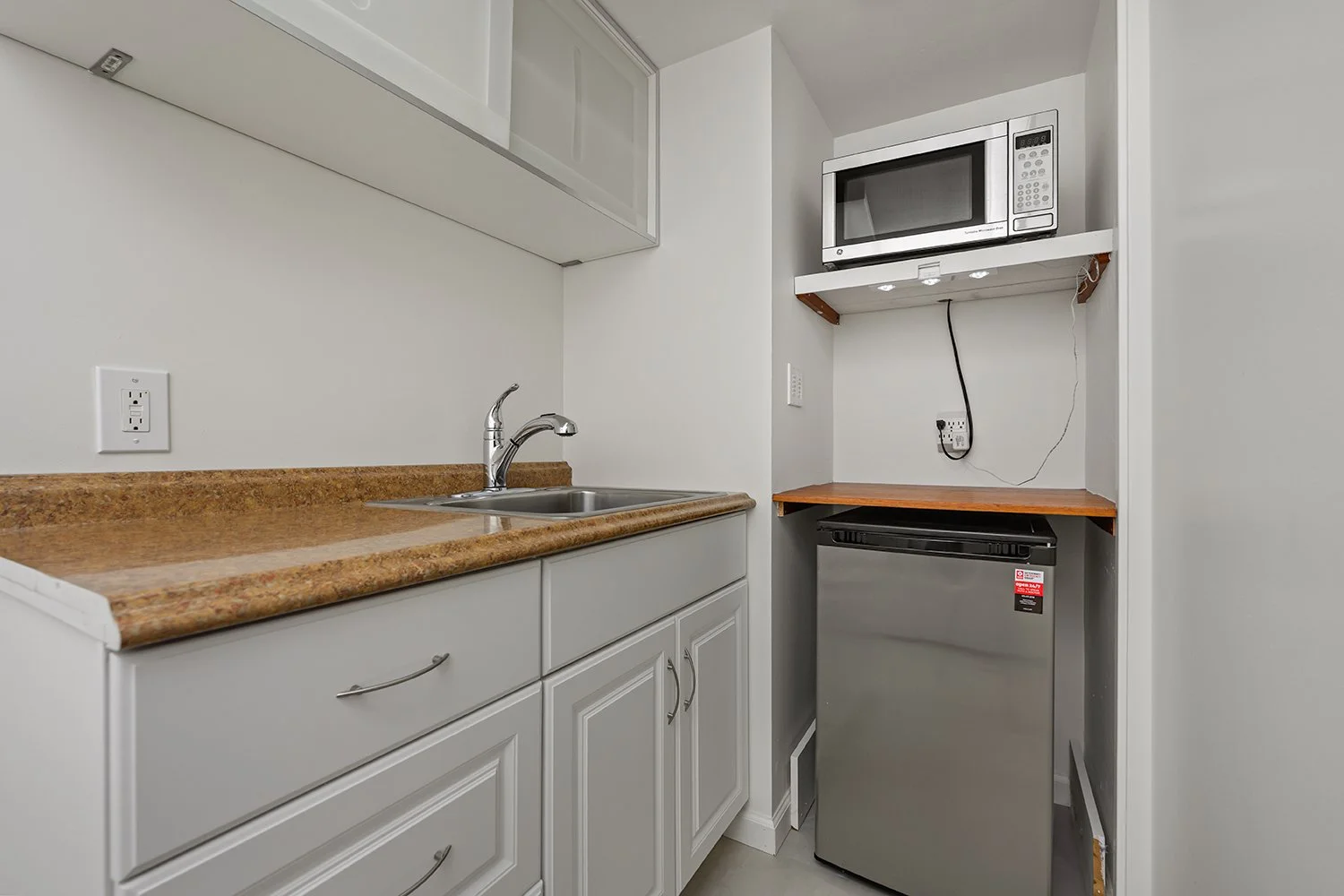 Small kitchen area with a white cabinet and a brown countertop, a sink with a faucet, a microwave oven on a shelf, and a small stainless steel refrigerator.