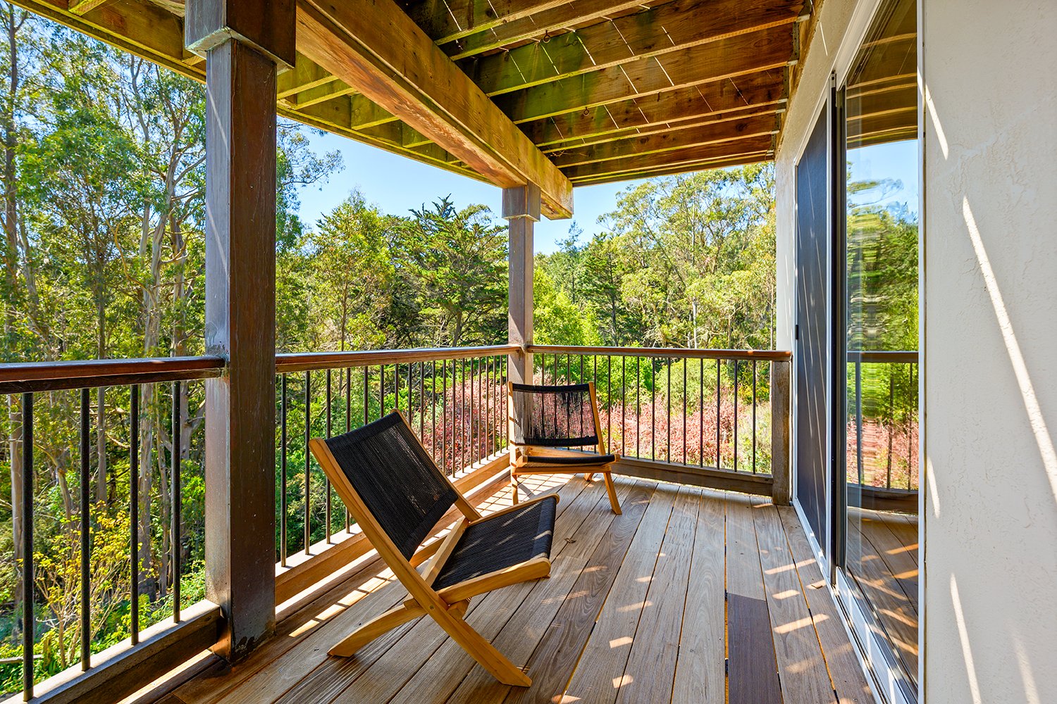 A wooden balcony with two black and wood chairs overlooking a green, leafy landscape with trees and a clear blue sky.