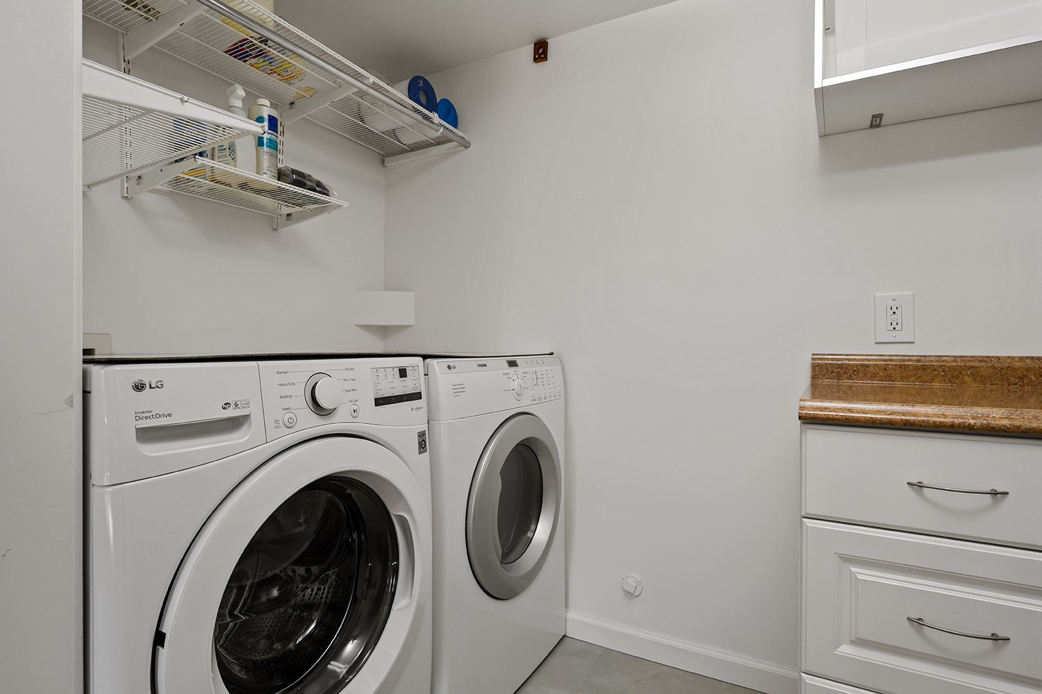 A laundry room with a front-loading LG washing machine and a top-loading white dryer, a granite countertop, white walls, and metal wire shelves holding cleaning supplies.